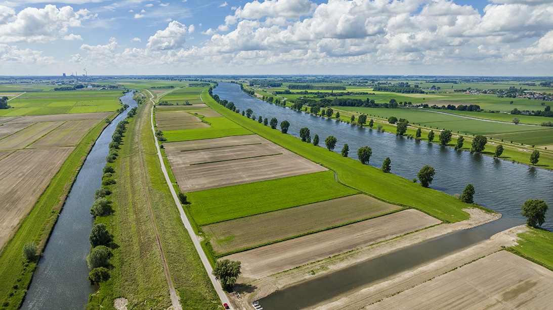Dronefoto van Capelsche uiterwaard met groen en bruin landschap met rechts de Maas en links een kanaaltje.